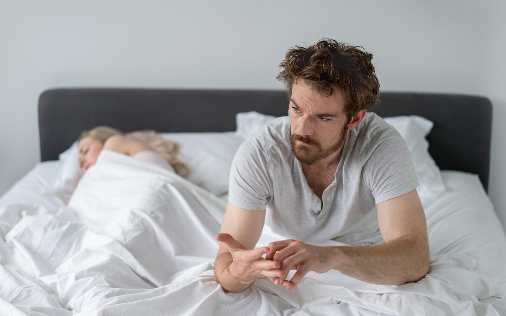 Mann sittende i sengen med en kvinne sovende ved siden av