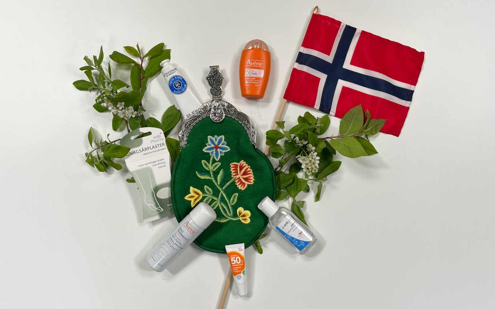 Grønn bunadsveske omgitt av ulike produkter som solkrem, antibac, gnagsårplaster og kildevannspray. Et norsk flagg og bjørkekvister.