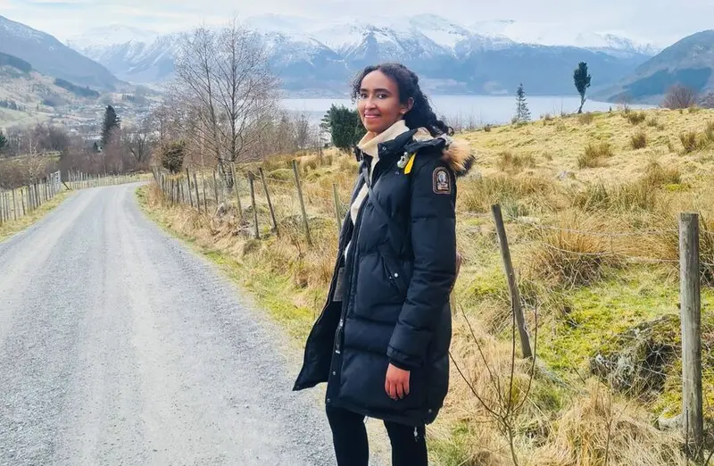 Bildet viser ambulerende farmasøyt Samiya gående langs en vei, mens hun smiler til kamera. I bakgrunnen ser man høye fjell og fjorder.