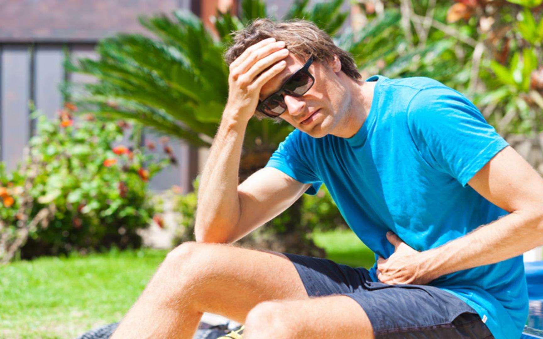 En mann som sitter i hagen, ene hånden holder seg på magen og ene hånden på pannen