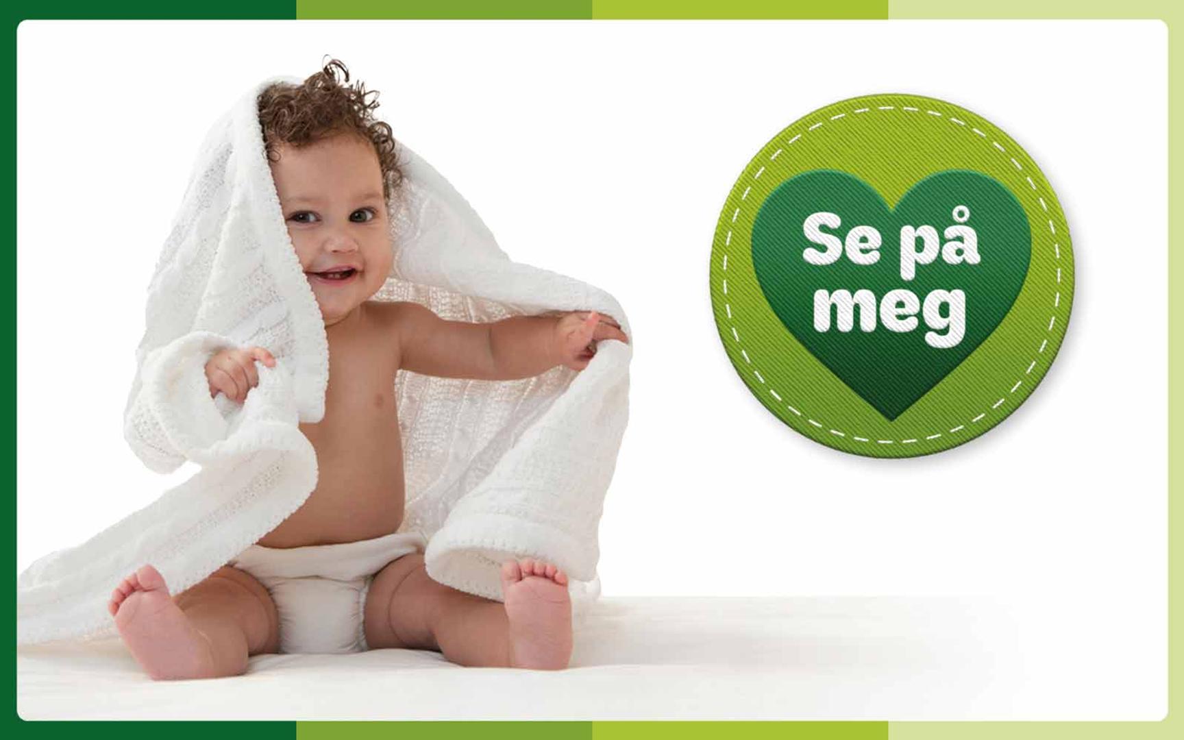 Smilende baby som sitter på gulvet med et hvitt teppe rundt seg, ved siden av en grønn hjerteformet badge med teksten 'Se på meg'.
