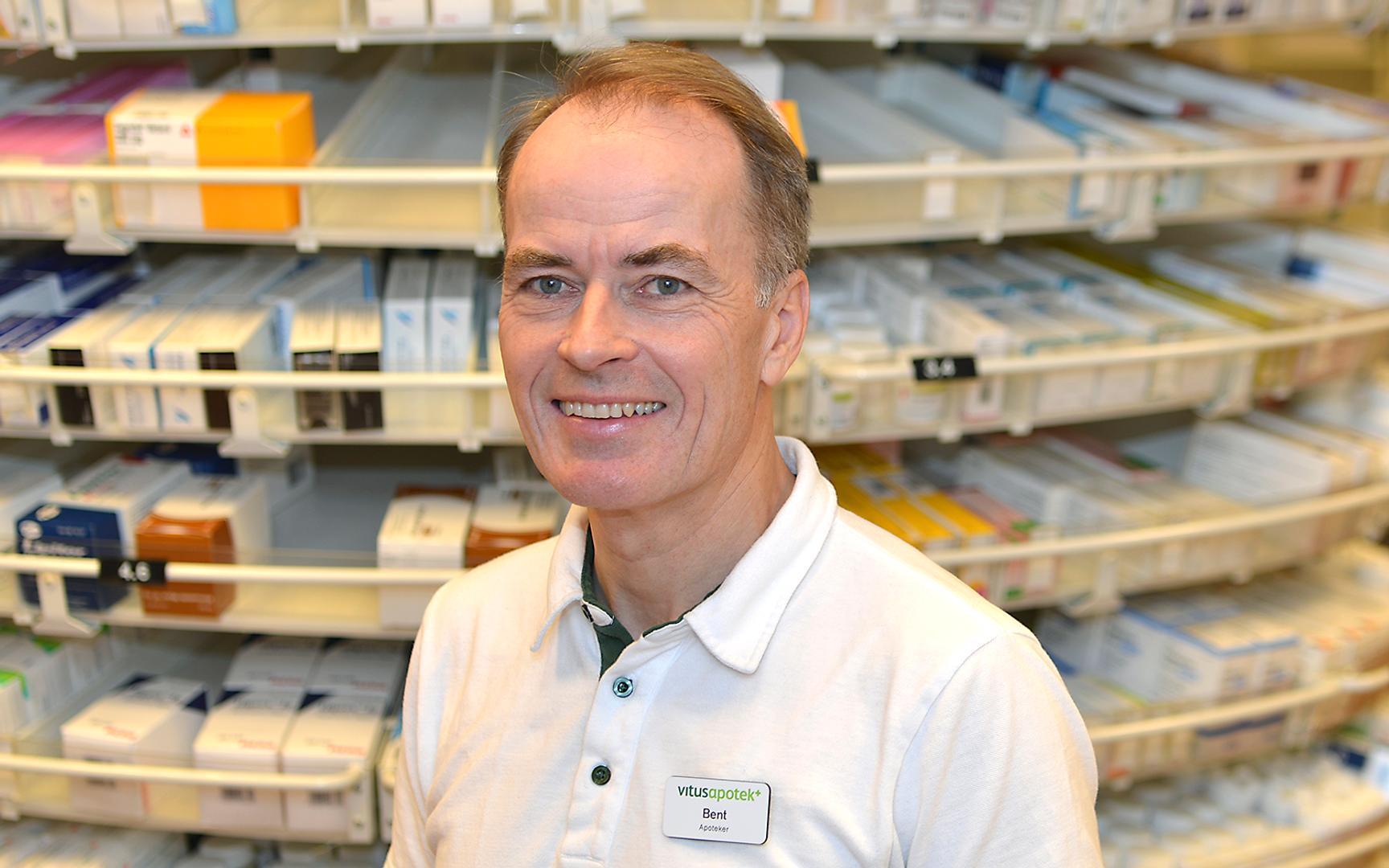 Mann som er farmasøyt som står foran medisinhyllen og smiler på bildet.