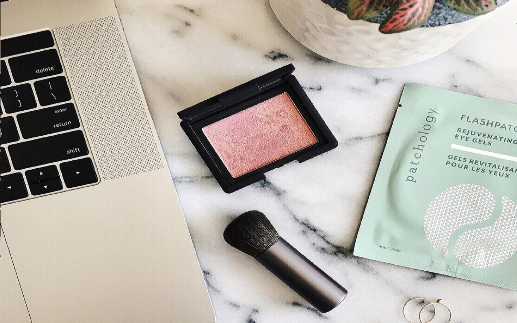 Flatlay-bilde av laptop på marmorbord sammen med blush, sminkekost, øyemasker og plante i bakgrunnen.