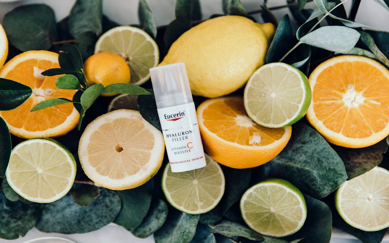 Bilde av appelsiner, sitron og lime på grønn blader. En flaske Eucerin ligger oppå fruktene