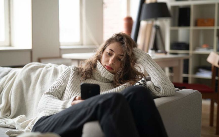 Bilde av en ung kvinnene sittende i sofaen med mobil i hånden. 
