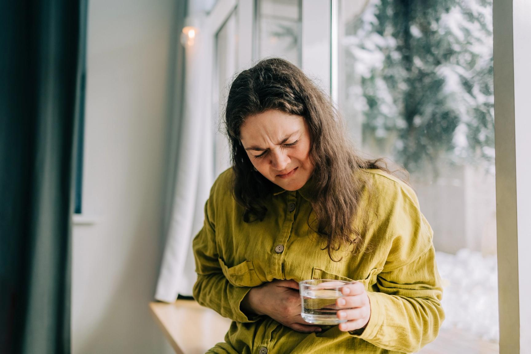 En person sitter ved et vindu og holder seg på magen med smerteuttrykk i ansiktet, mens de holder et glass vann i den andre hånden.