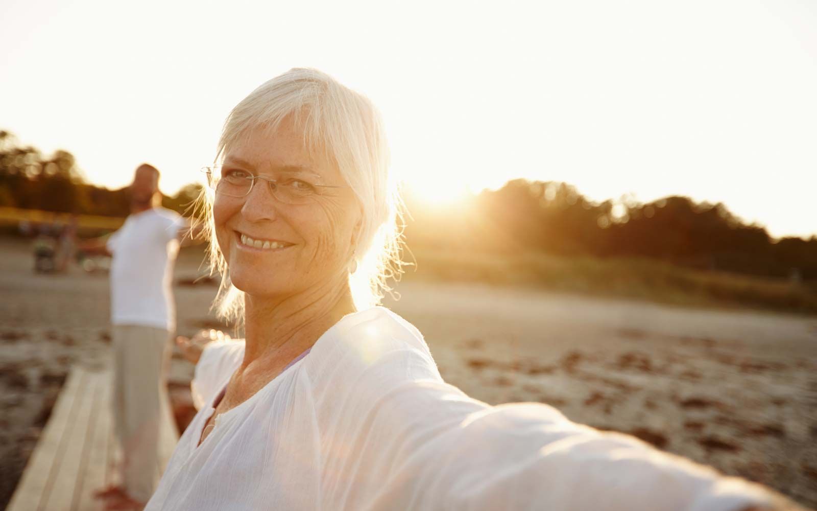 En kvinne tar en selfie på en strand med en mann i bakgrunnen