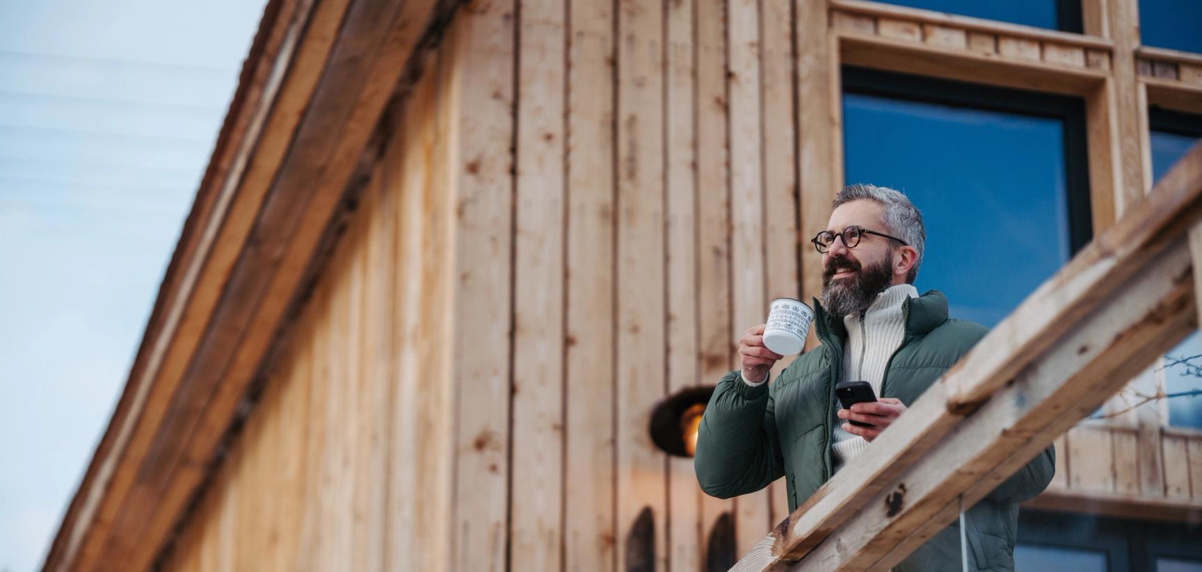 Mann med kaffekopp og mobil i hånden stående på en veranda ved en hytte.