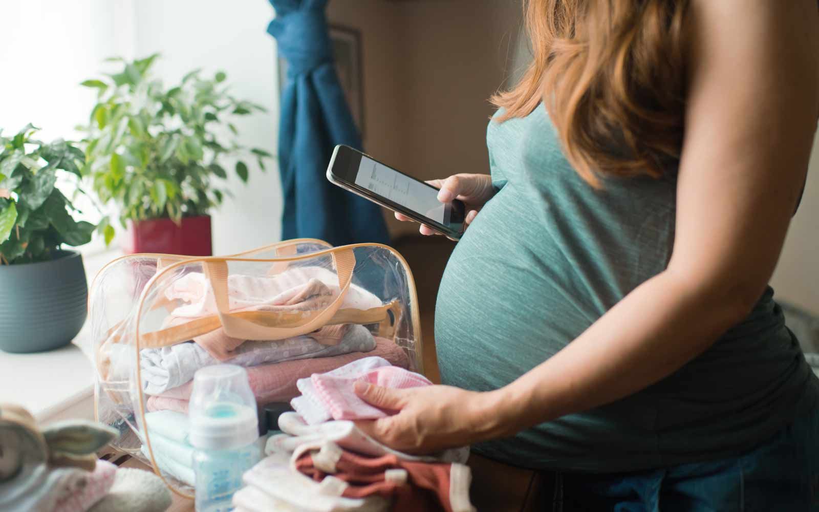 Gravid kvinne ser ned på en liste på mobilen sin samtidig som hun pakker klær og utstyr til fødebagen.