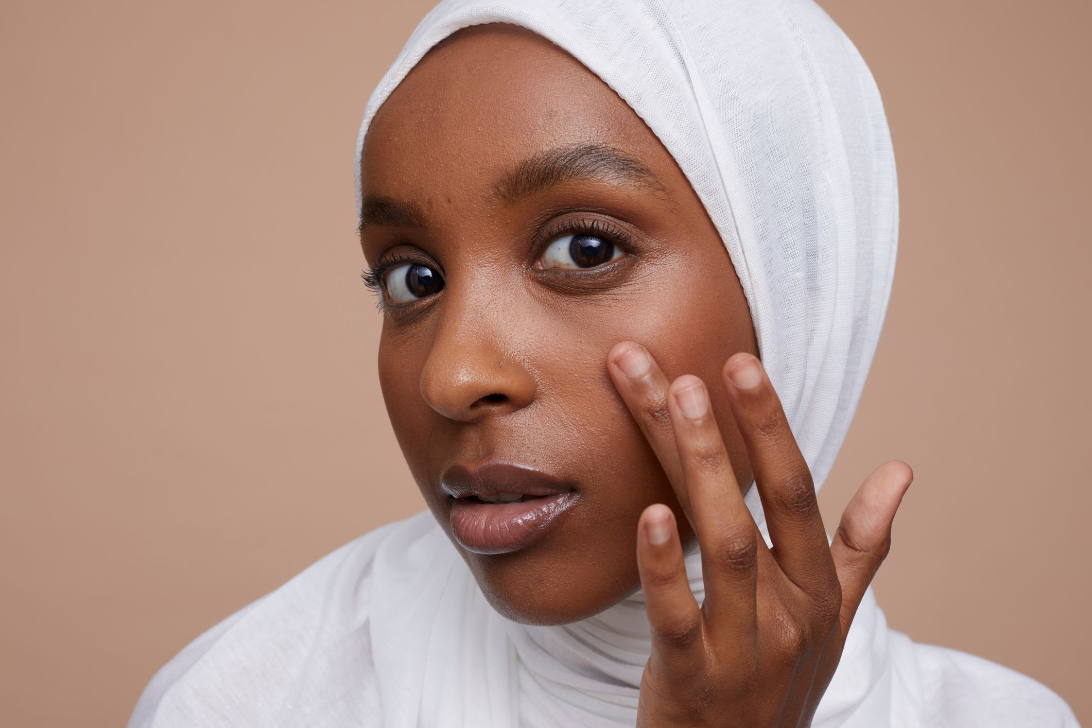 En ung kvinne iført en hvit hijab ser på kameraet mens hun forsiktig berører ansiktet sitt. Huden hennes ser sunn ut, og hun ser ut til å være i ferd med å bruke hudpleieprodukter. Bakgrunnen er nøytral, med en myk beige farge som fremhever hennes naturlige skjønnhet.