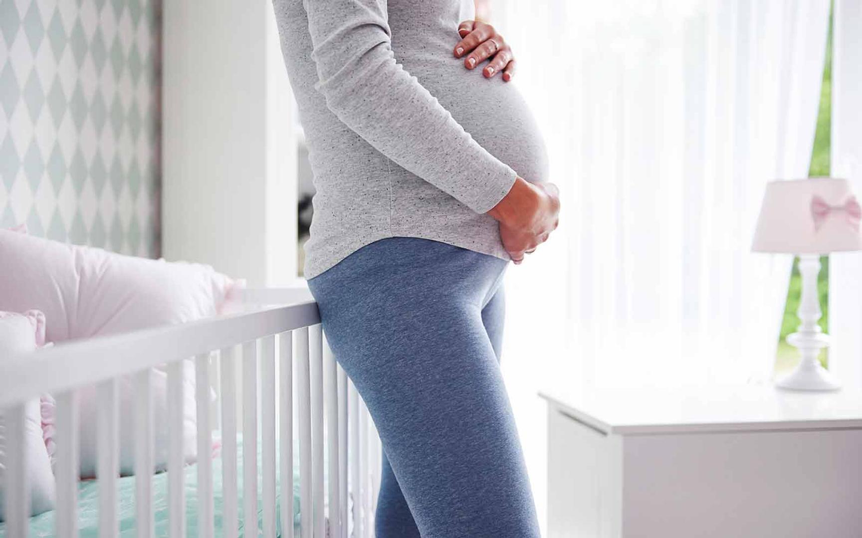 Bilde av en gravid dame som holder hendene over magen, stående ved en barneseng.