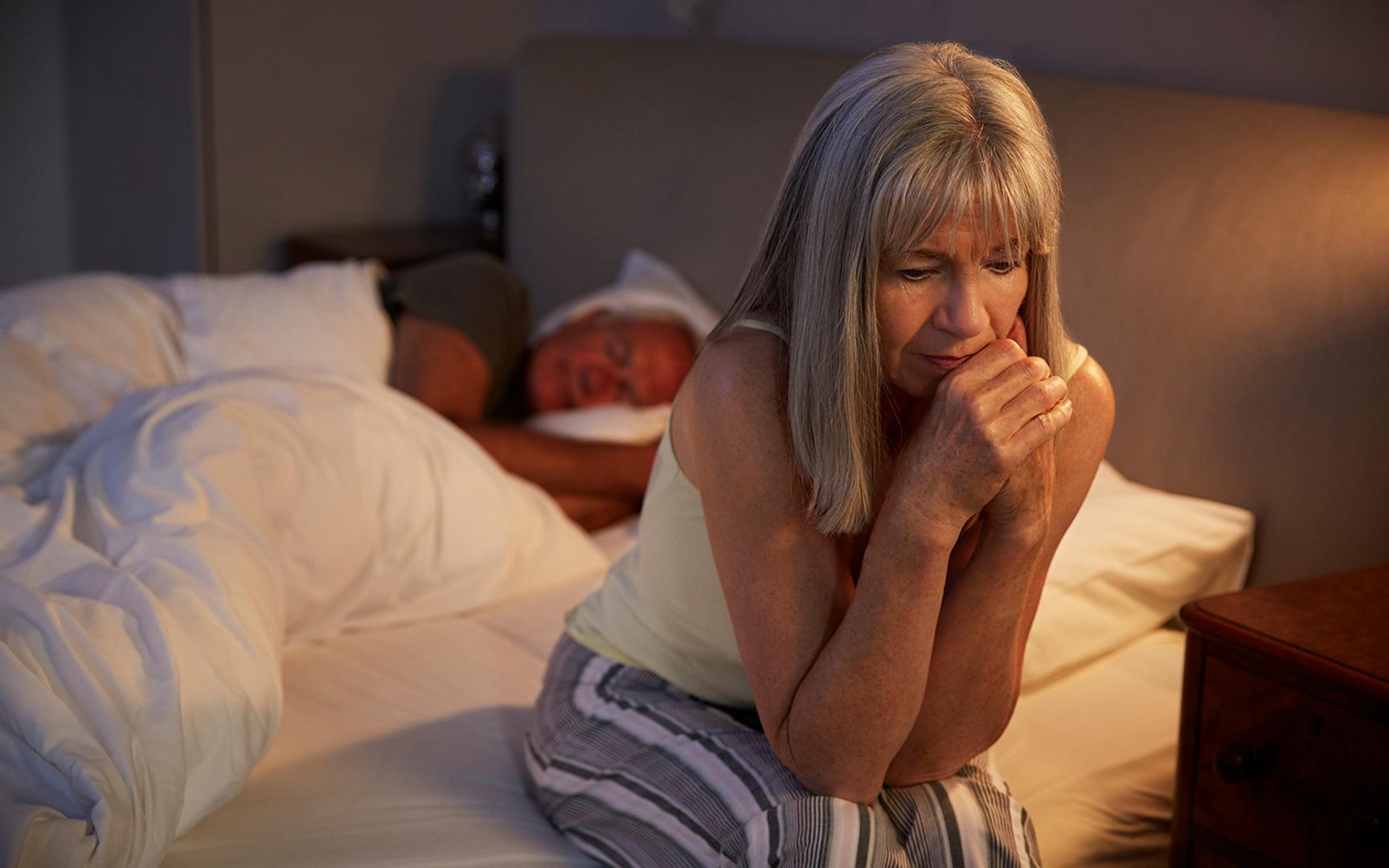 Kvinne sittende på sengekanten og en mann sovende i sengen bak.