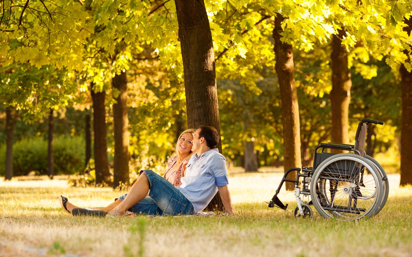 En kvinne og en mann sitter på plenen i park med en tom rullestol ved siden av. 