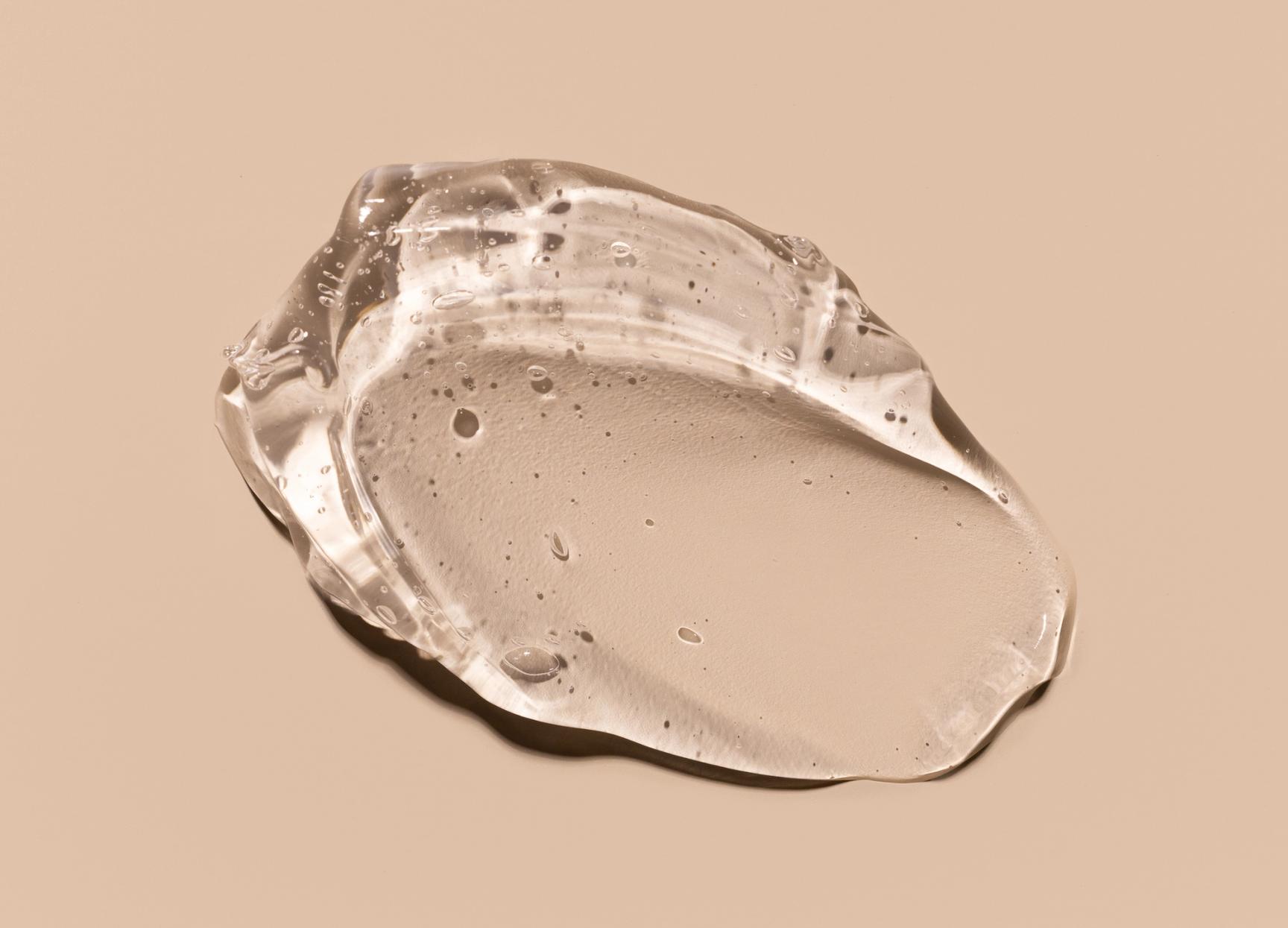 Gjennomsiktig, geléaktig hudpleieprodukt med små luftbobler, plassert på en lys beige bakgrunn. Teksturen ser fuktighetsgivende og glatt ut.