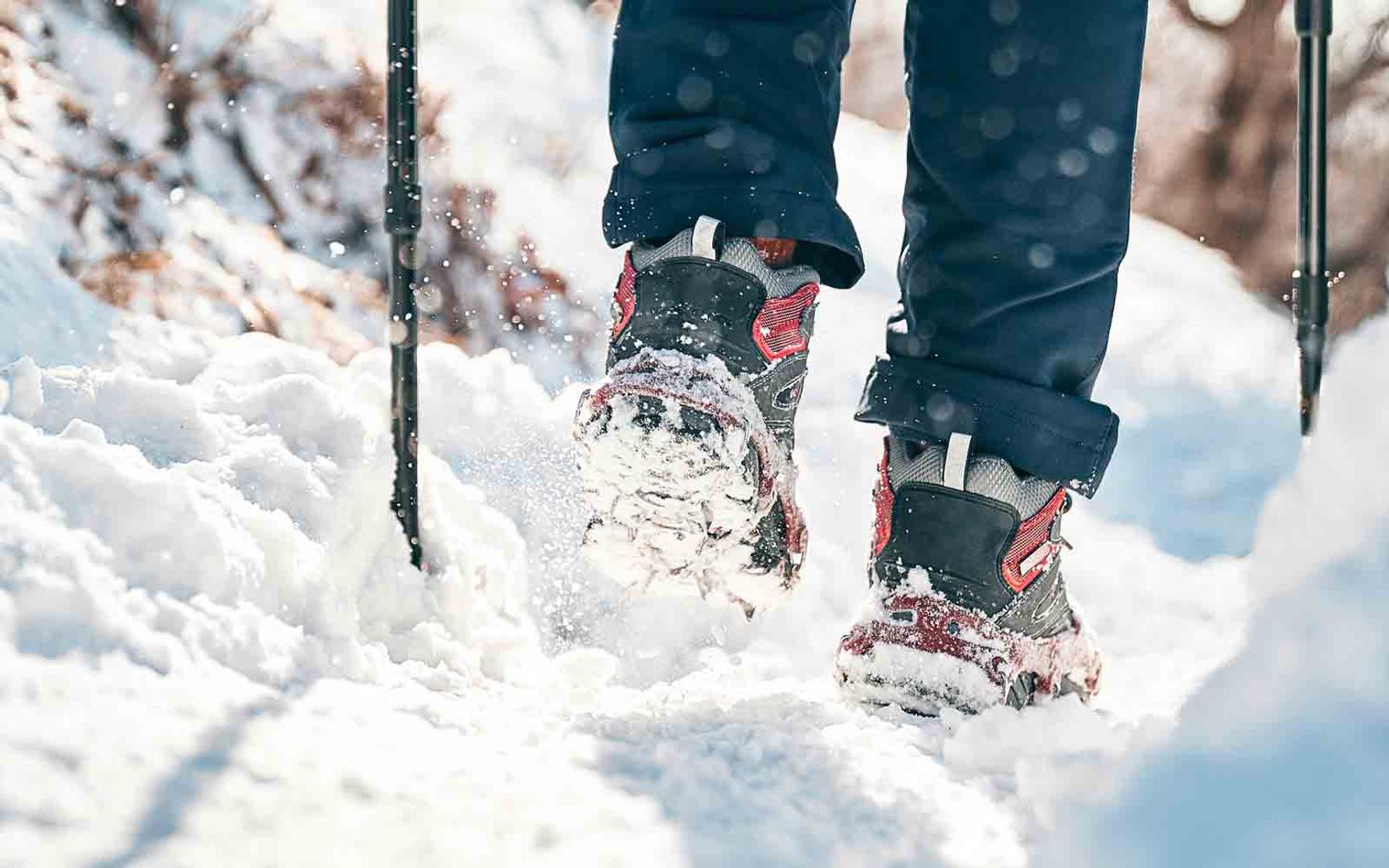 Person med vintersko som går i løs snø i skogsterreng. Bildet er tatt bakfra med fokus på skoene, som har brodder på.