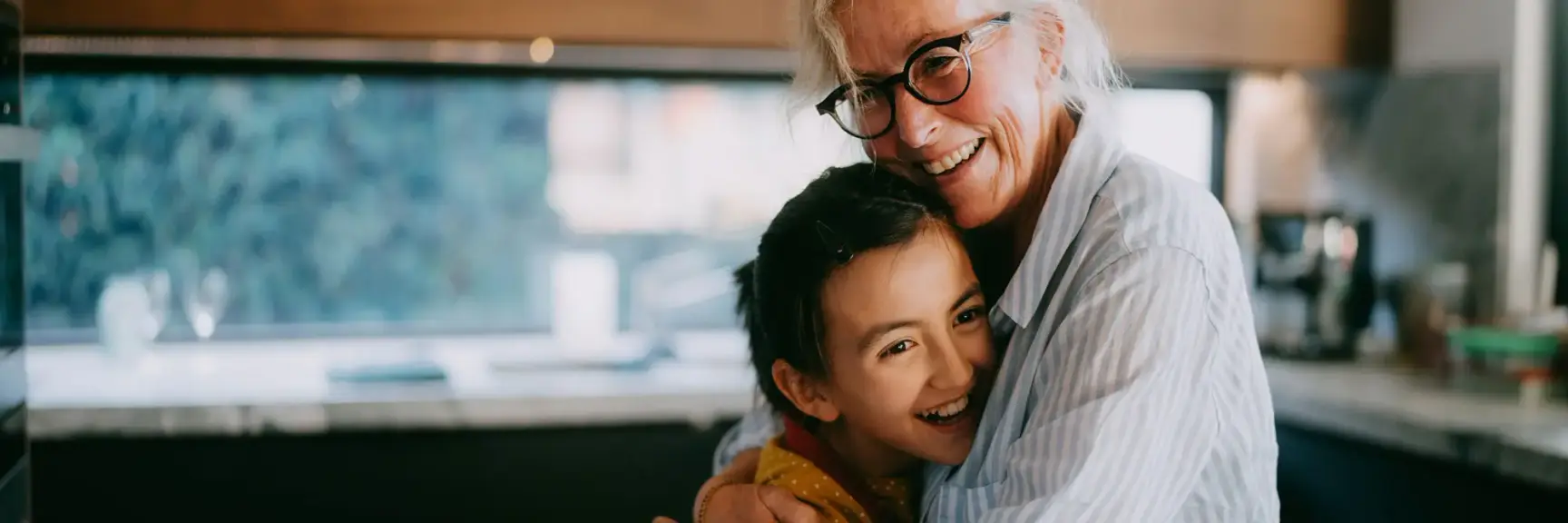 Bestemor holder rundt og klemmer barnebarnet sitt på et kjøkken, i et varmt lys