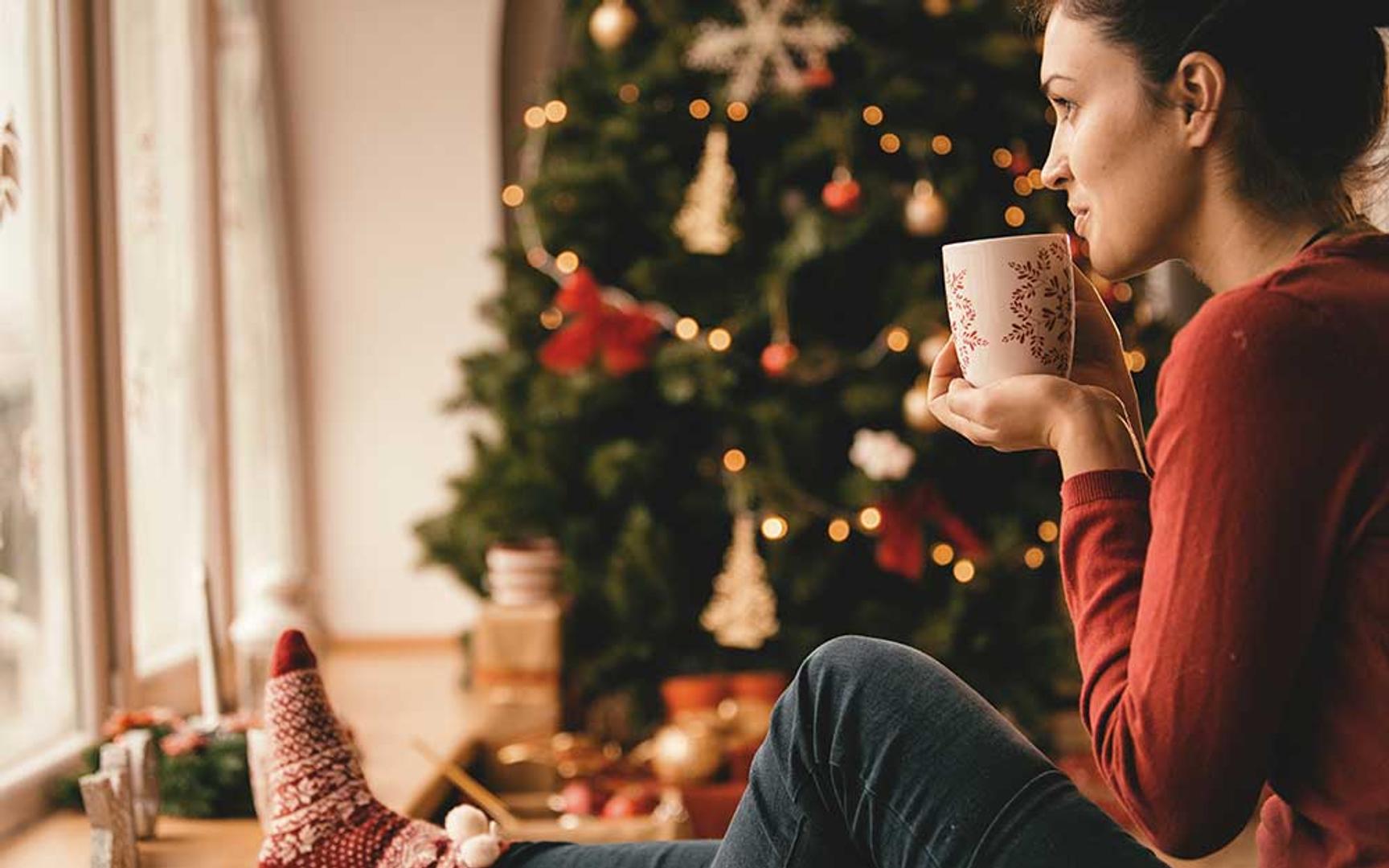 En dame som sitter på gulvet med en kopp mot ansiktet og et juletre i bakgrunnen