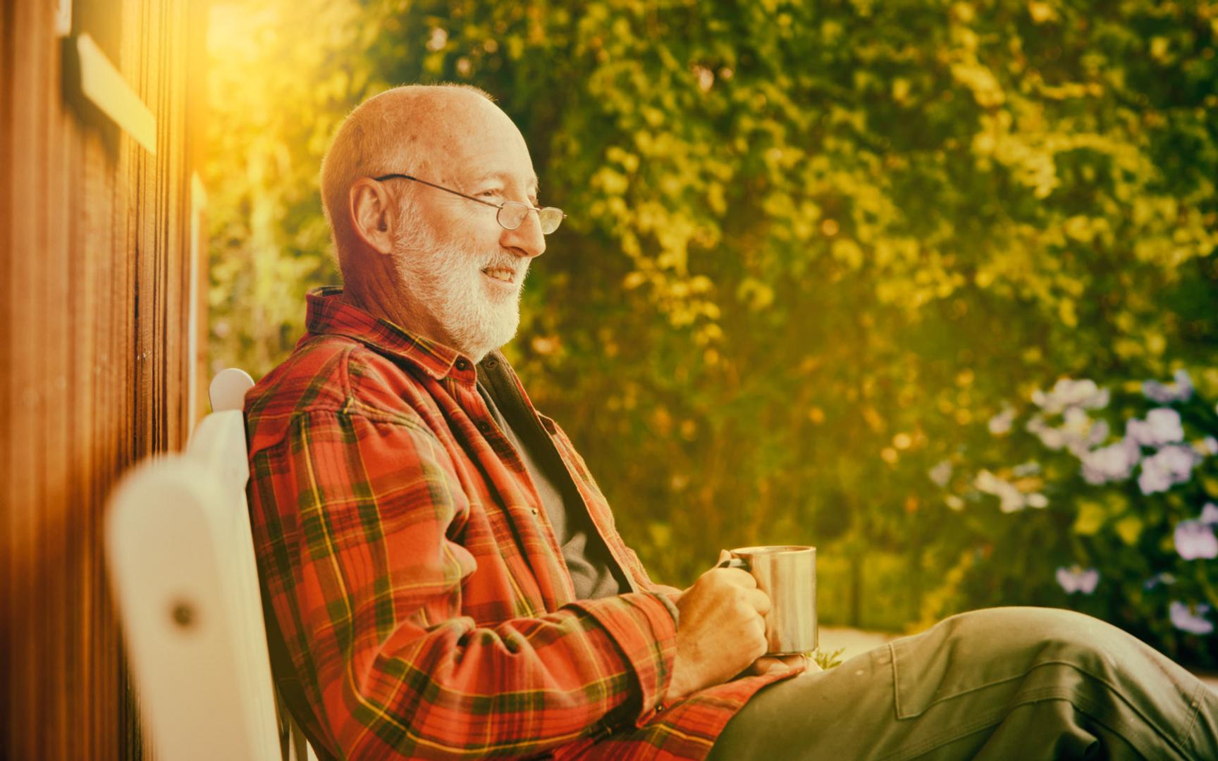 Bilde av en eldre mann sittende på en stol i husveggen med en kopp i hånden.