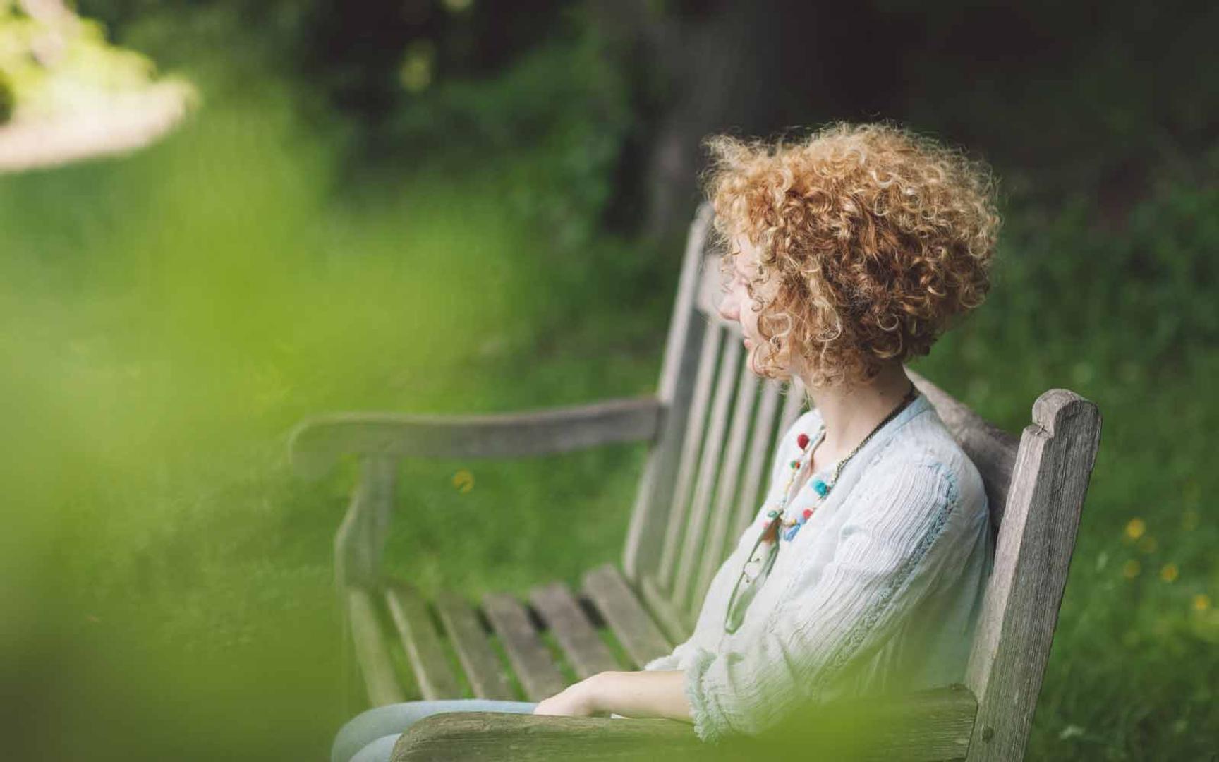 En kvinne med krøllete hår sitter på en park benk med ansiktet vendt bort fra kameraet