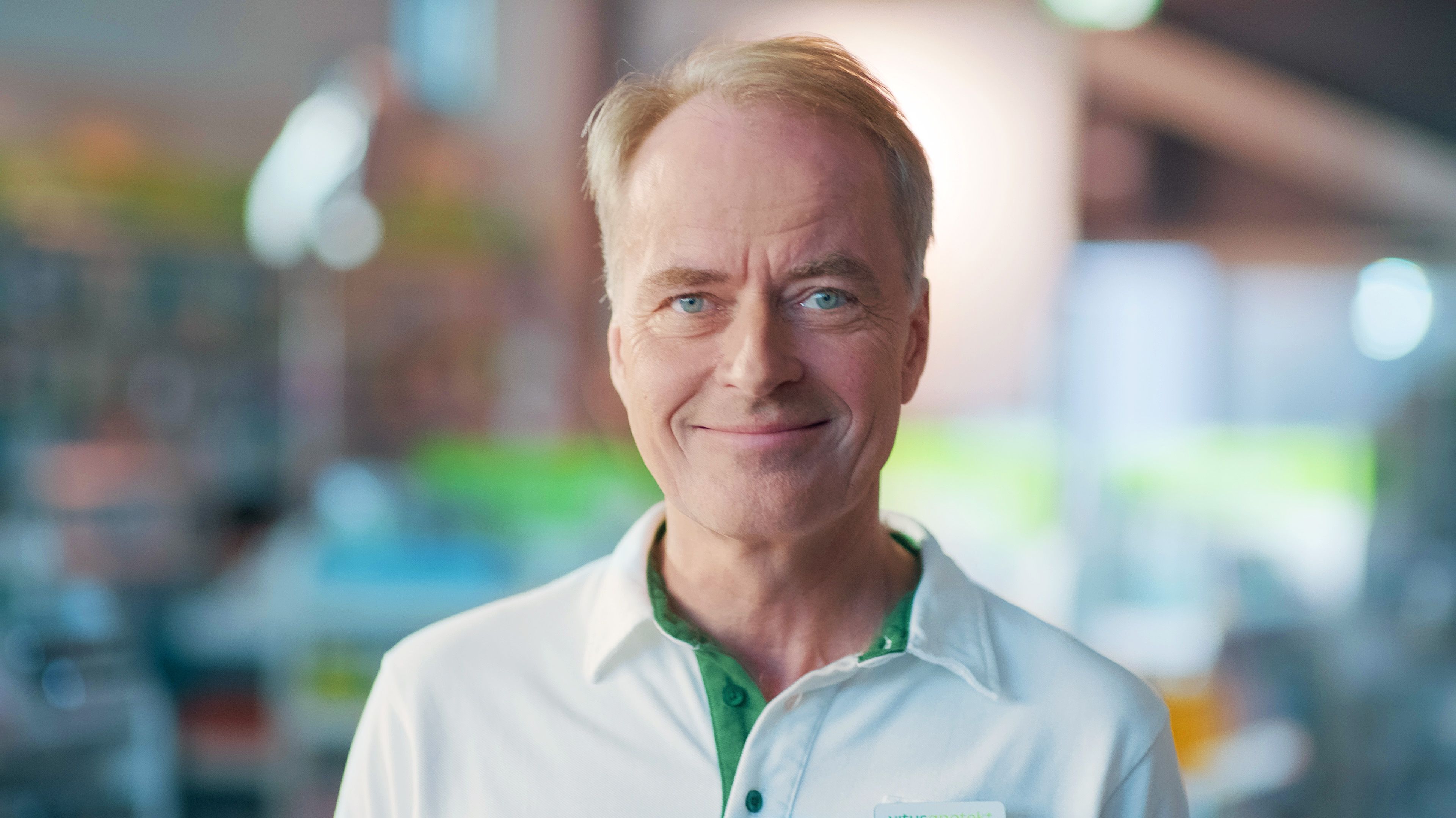 Nærbilde av ansiktet til en farmasøyt med uniform som ser i kamera og smiler lunt, bakgrunnen er i ufokus