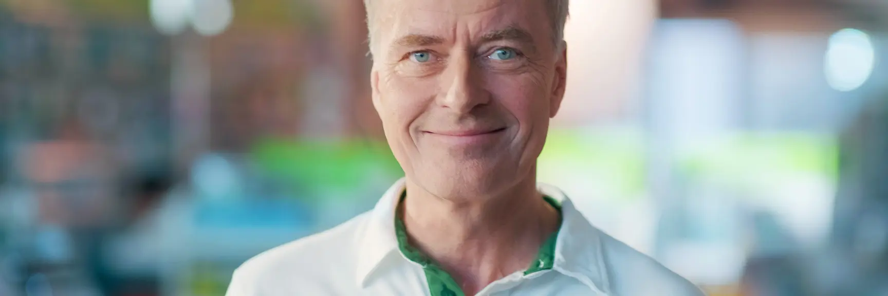 Nærbilde av ansiktet til en farmasøyt med uniform som ser i kamera og smiler lunt, bakgrunnen er i ufokus