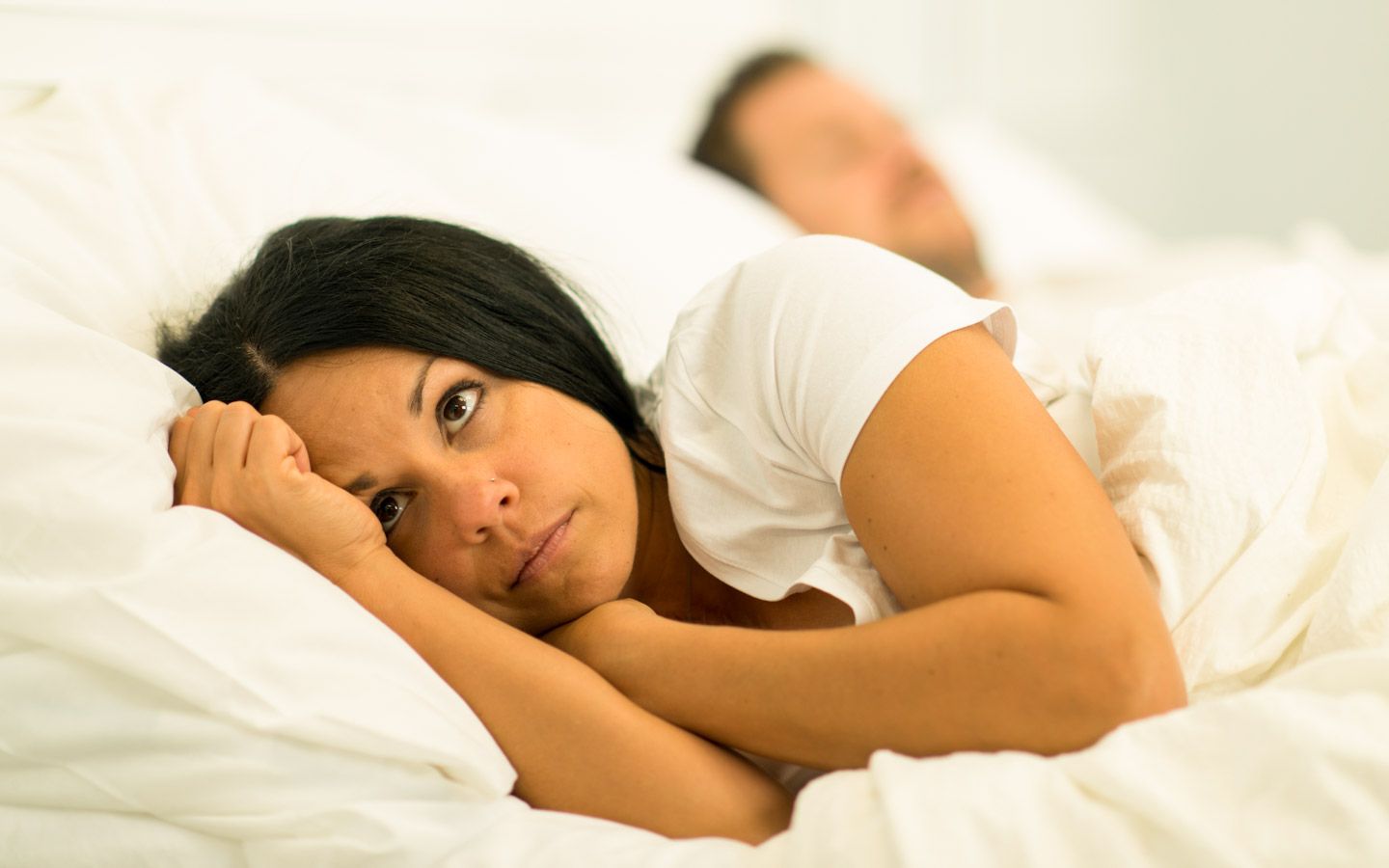 Kvinne som ligger våken i en seng. Hun ser tankefull eller bekymret ut. Bak henne ligger en mann og sover.
