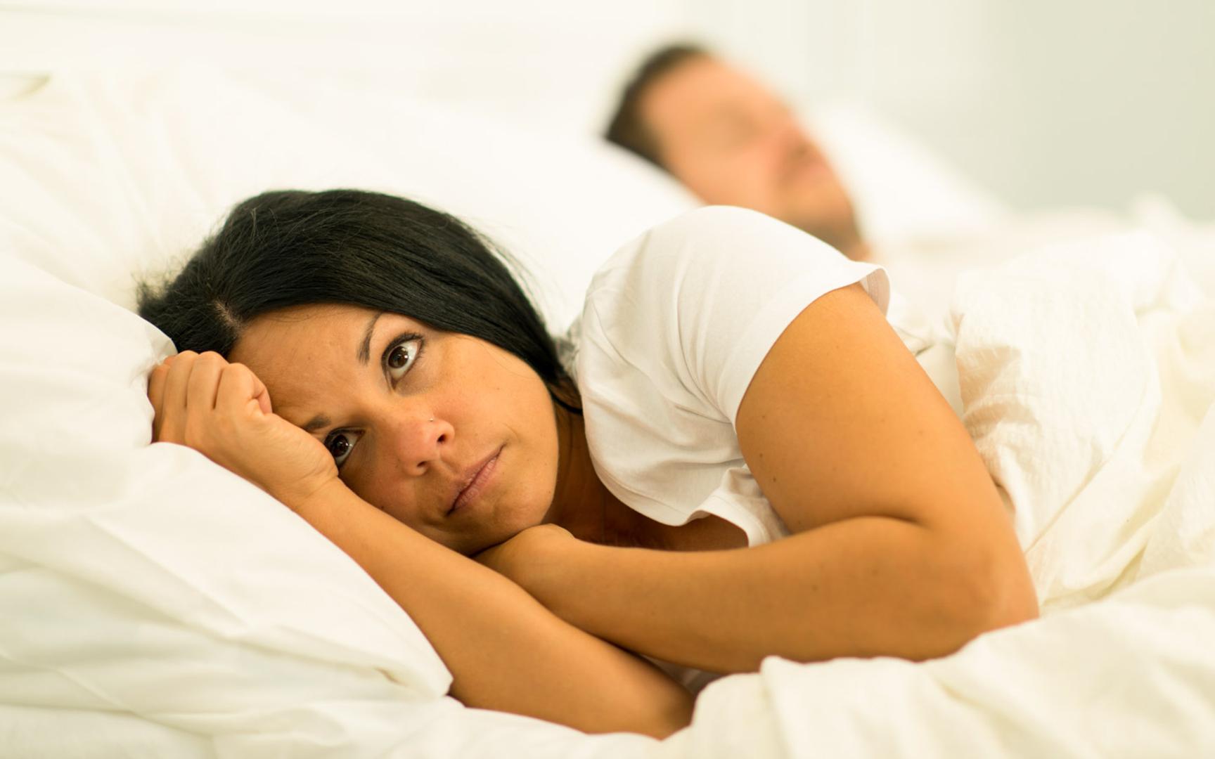 Kvinne som ligger våken i en seng. Hun ser tankefull eller bekymret ut. Bak henne ligger en mann og sover.
