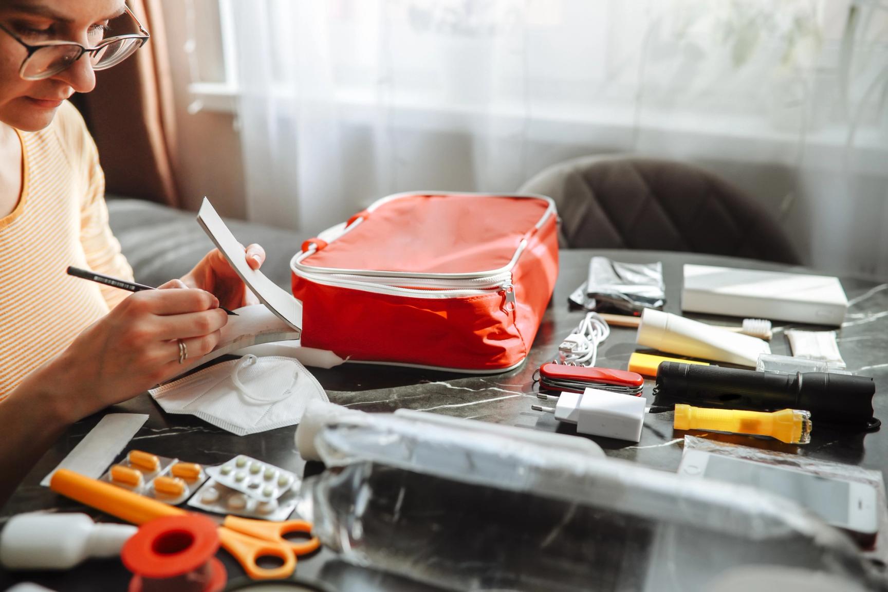 En person sitter ved et bord og pakker et rødt førstehjelps- eller reiseapotek. Rundt henne ligger ulike nødvendige artikler som medisiner, plaster, saks, ansiktsmasker, lommelykter, powerbank, ladere og annet nødutstyr