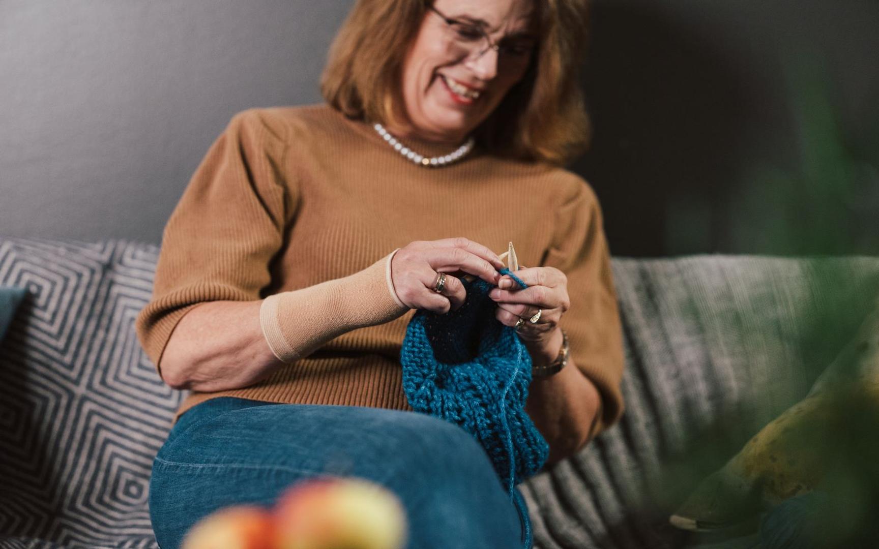 En kvinne sitter i en sofa og strikker med håndleddsstøtte