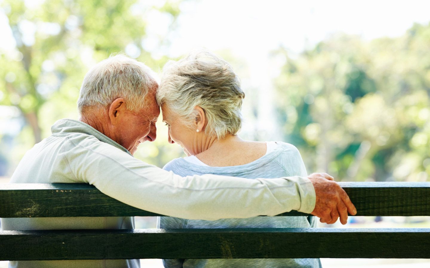 Bilde av et eldre par bakfra sittende på en benk, mannen holder rundt kvinnen. 