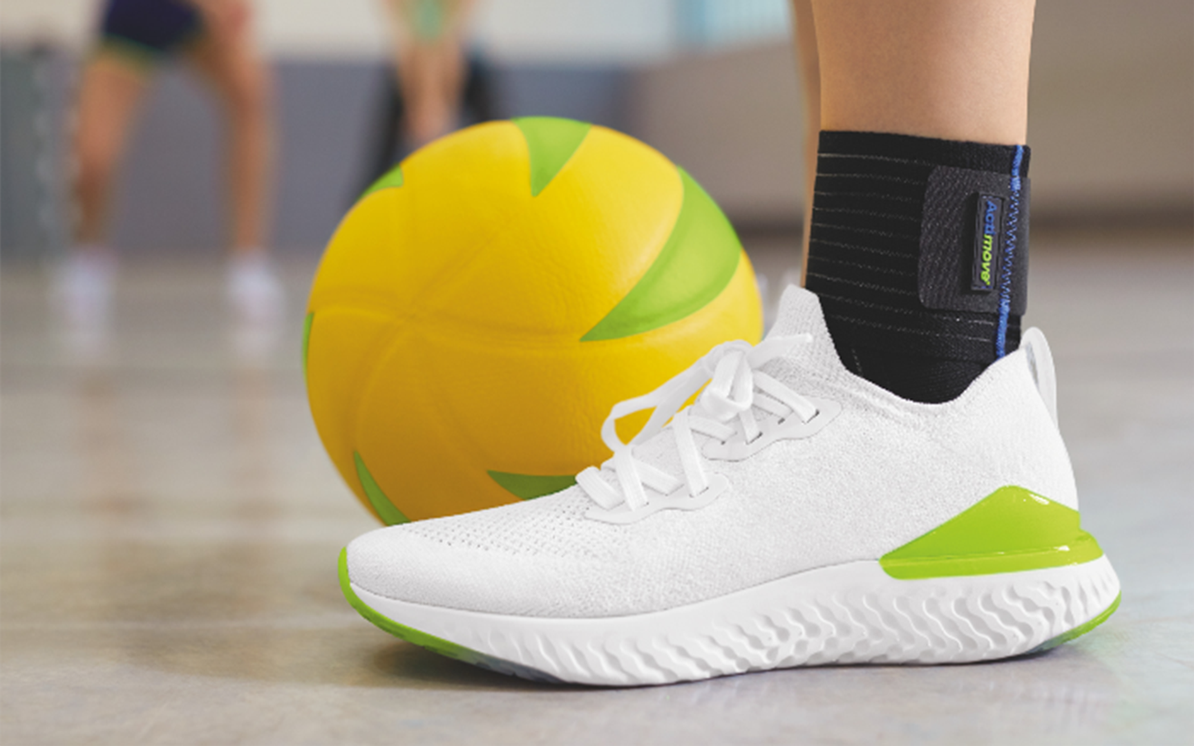 Person med svart ankelstøtte og hvit treningssko står ved siden av gul ball i gymsal