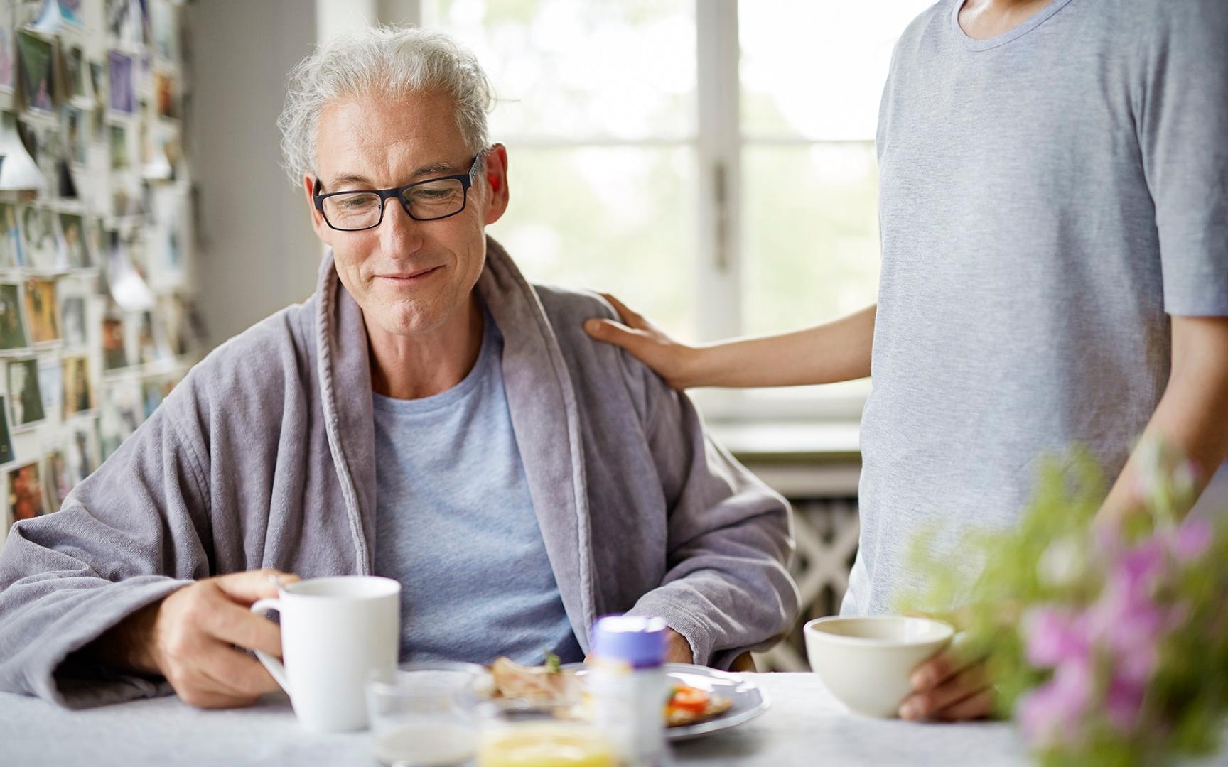 En mann sitter ved et bord med mat og noen holder han på skulderen