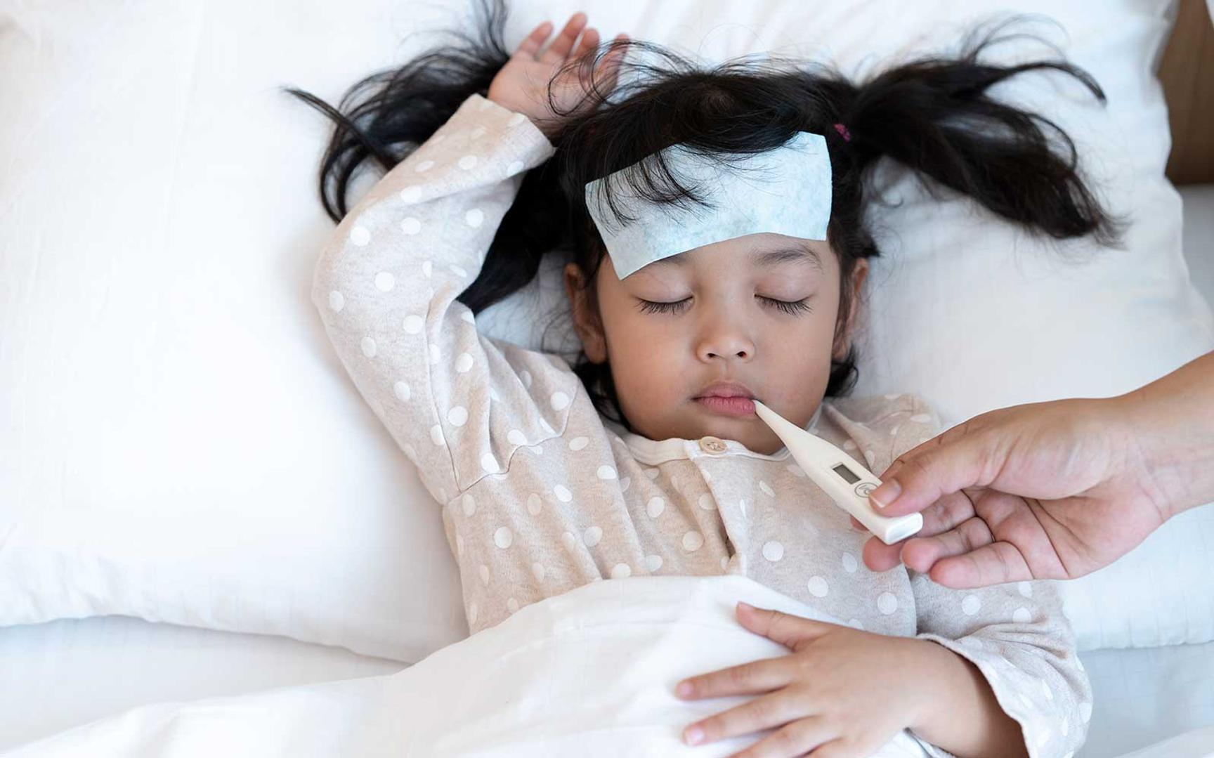 Et lite barn ligger på en seng med et febertermometer i munnen. Feber kan behandles med smertestillende og febernedsettende legemidler.