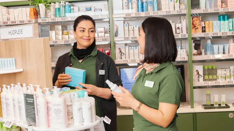 To farmasøyter i ett apotek smiler og ser på hverandre, i bakgrunnen skimtes hyller med varer.
