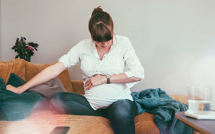 En gravid kvinne sitter på sofaen og holder seg på magen, trolig fordi riene har startet.