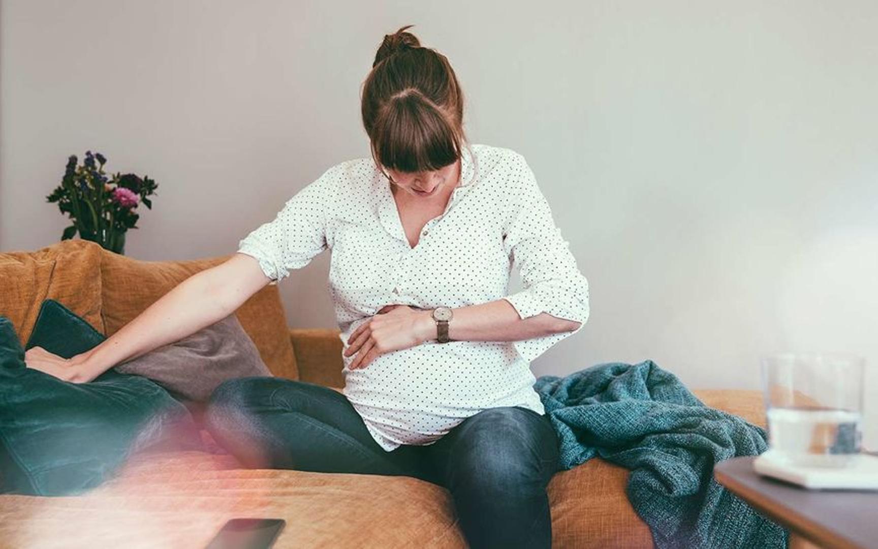 En gravid kvinne sitter på sofaen og holder seg på magen, trolig fordi riene har startet.