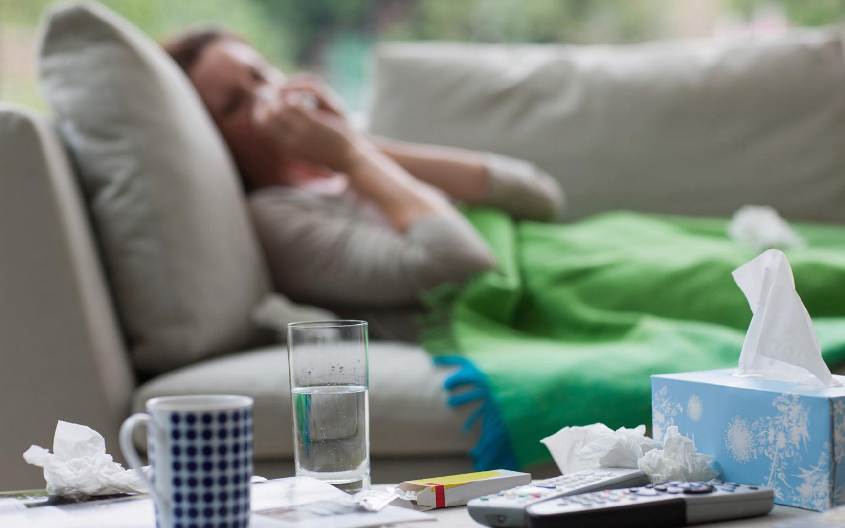 Bilde av en dame som snyter seg liggende på sofaen med pledd, på bordet står det vann, medisin, tørkepapir og fjernkontroller.