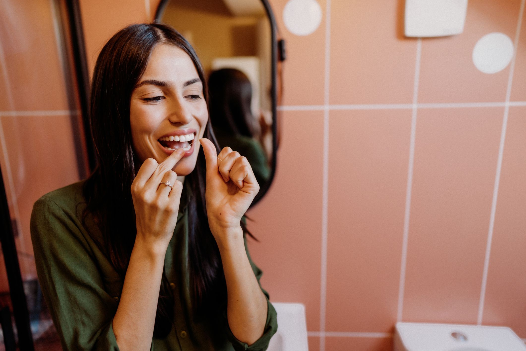 En kvinne med mørkt hår og grønn genser står på et bad og bruker tanntråd