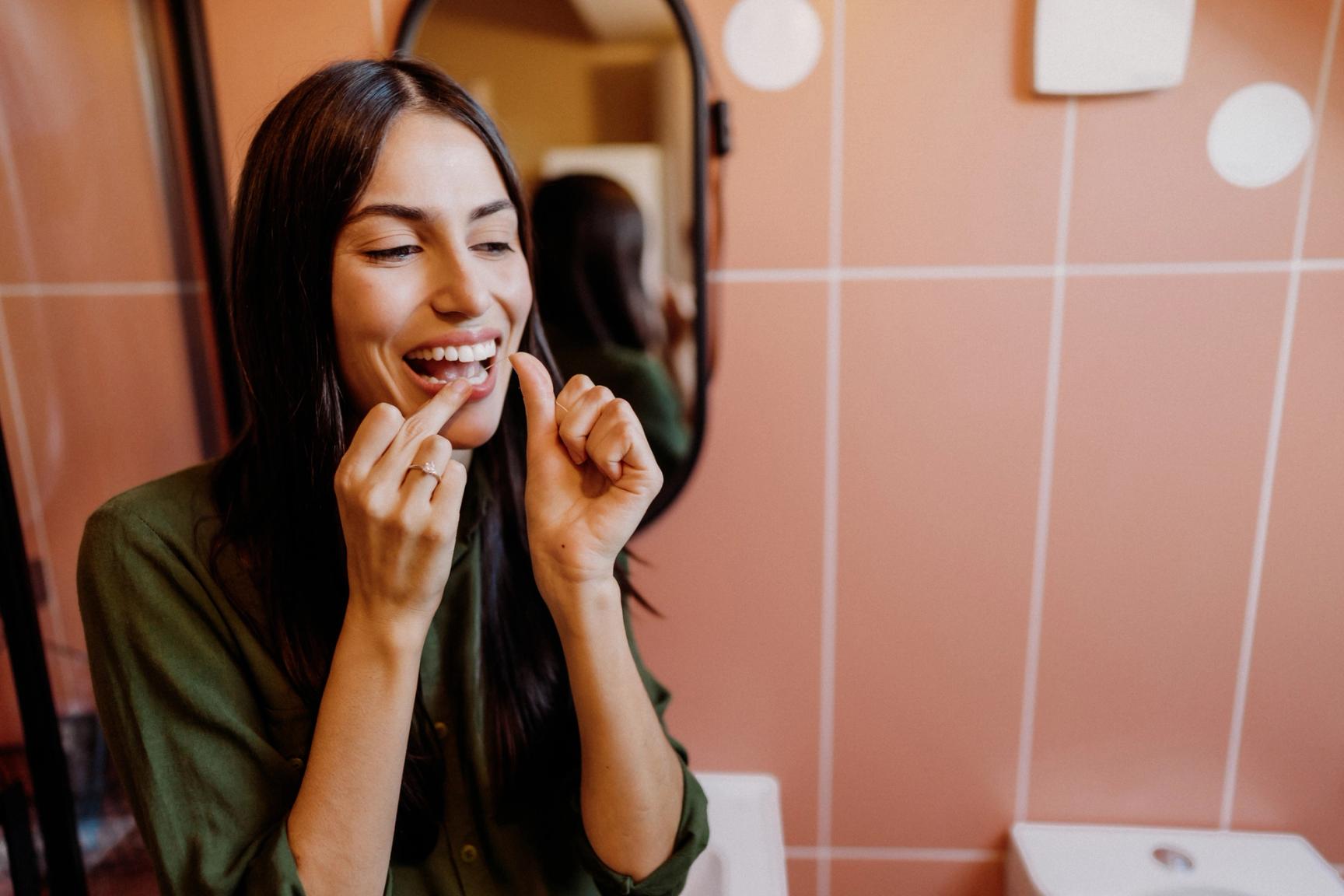 En kvinne med mørkt hår og grønn genser står på et bad og bruker tanntråd