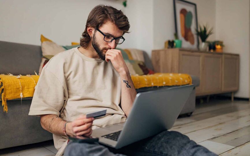 Bilder viser en tankefull mann som sitter på gulvet foran sofaen med en laptop i fanget og et kredittkort i hånden.