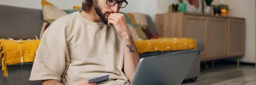 Bilder viser en tankefull mann som sitter på gulvet foran sofaen med en laptop i fanget og et kredittkort i hånden.