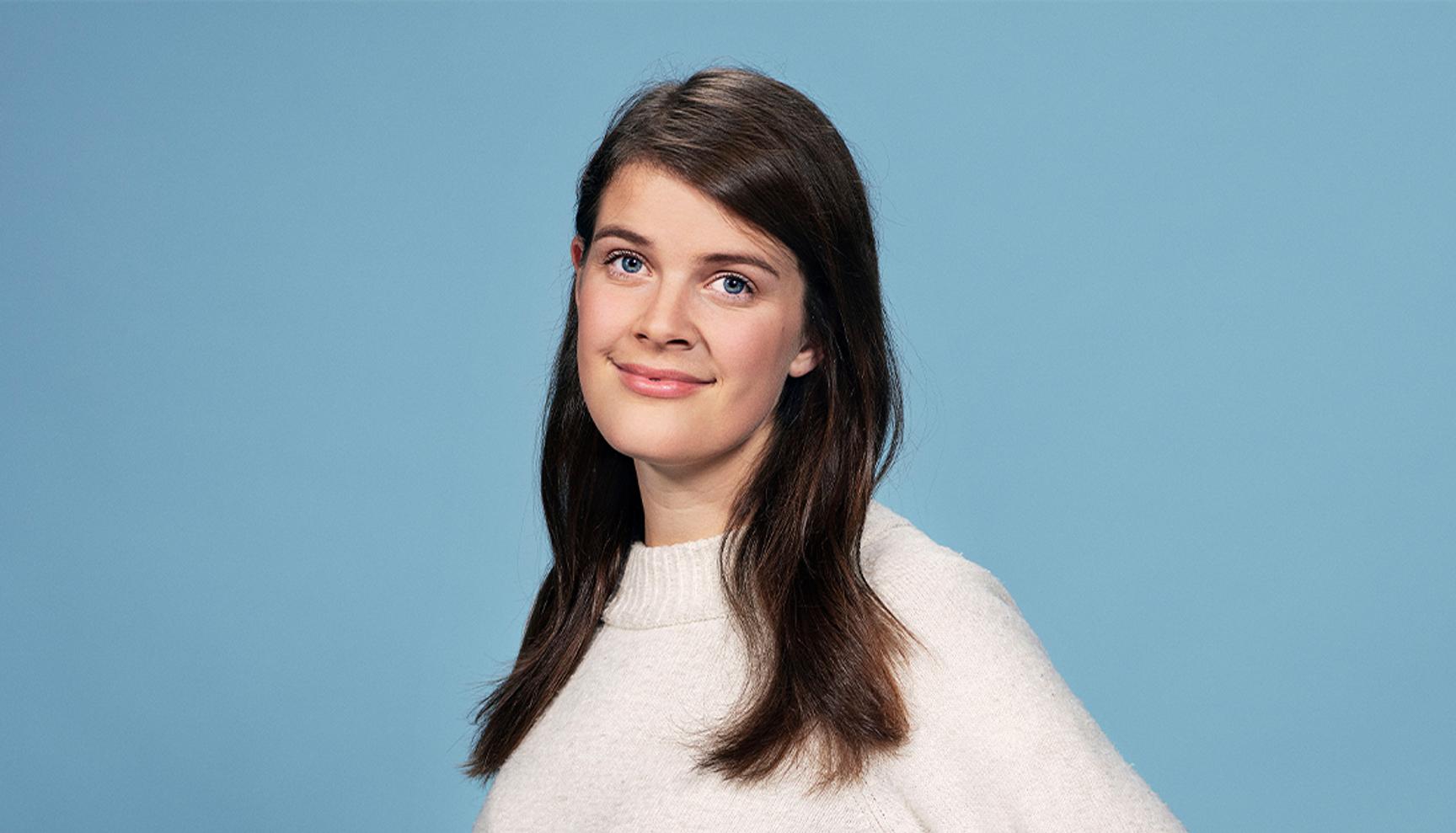 En kvinne med langt, brunt hår stor mot en lys blå bakgrunn, iført en lys genser og et vennlig uttrykk.