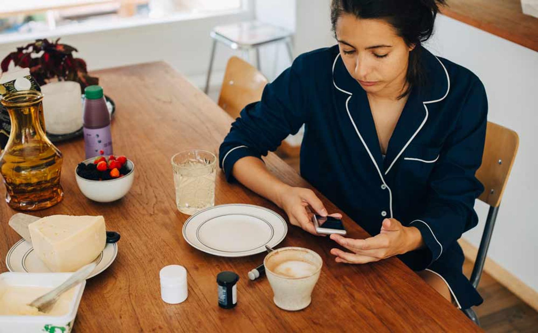 En dame som sitter ved frokostbordet med mobilen i hånden. På bordet er det kaffe, ost,bær og medisinglass.