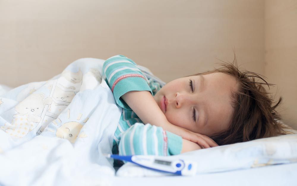 Bilde av et barn som sover i en seng med hendene under hodet og en febermåler ved siden av puten. 