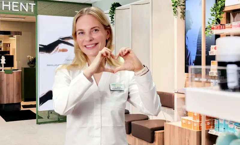 Farmasøyt smiler og lager et hjerte med hendene, man ser et apotek i bakgrunnen