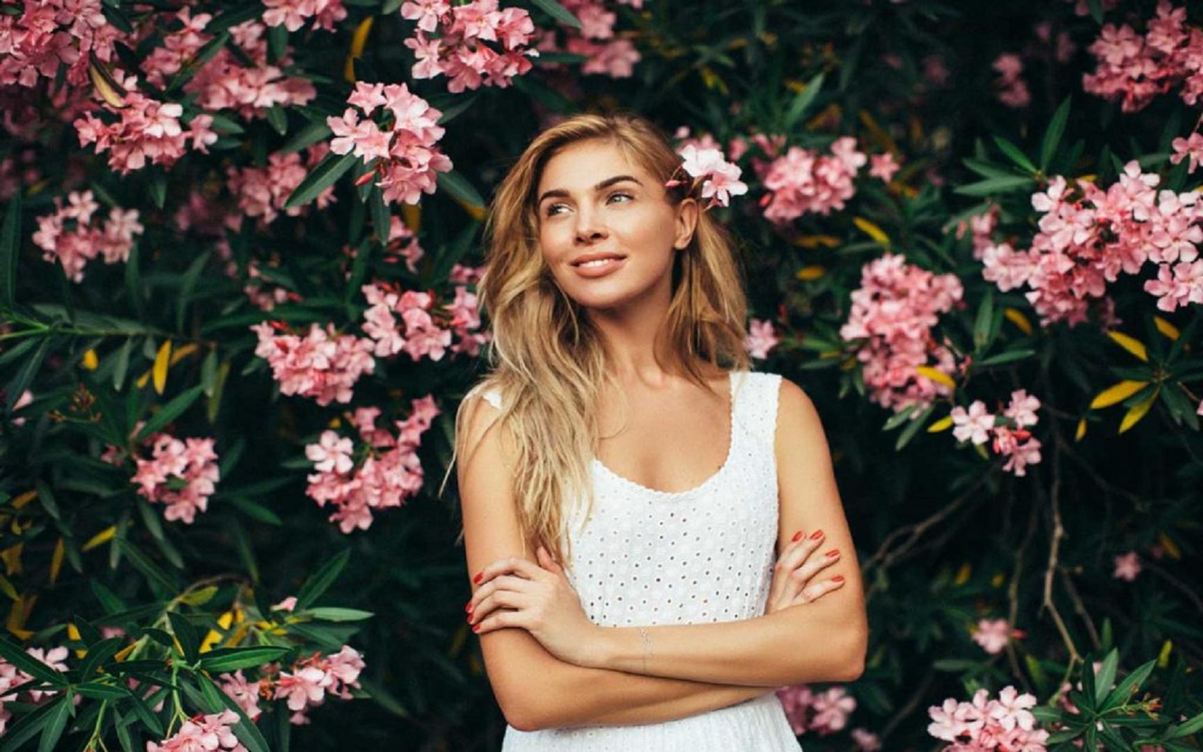 En dame med langt hår, hvit singlet og smiler utover med armene i kors. I bakgrunnen en grønn busk med rosa blomster.