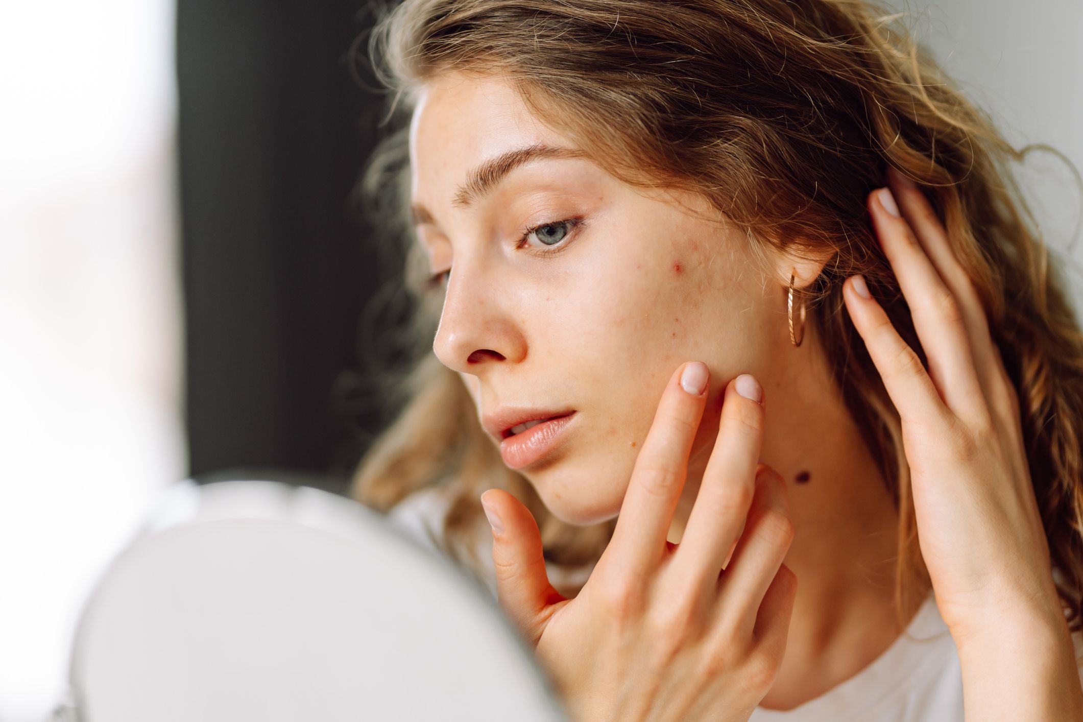 Ung kvinne ser på huden sin i et speil og berører kinnet, med fokus på hudpleie og dehydrert hud