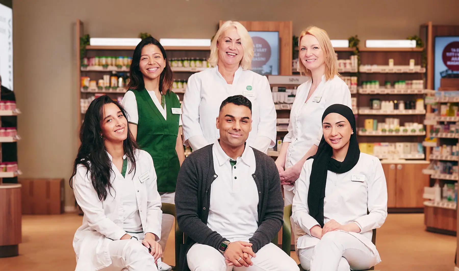 Gruppebilde av 6 ansatte i Vitusapotek i uniform som smiler og ser i kamera, bildet er tatt inne på et apotek og man ser hyllevarer i bakgrunnen. 