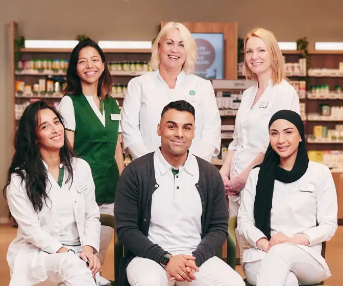 Gruppebilde av 6 ansatte i Vitusapotek i uniform som smiler og ser i kamera, bildet er tatt inne på et apotek og man ser hyllevarer i bakgrunnen.