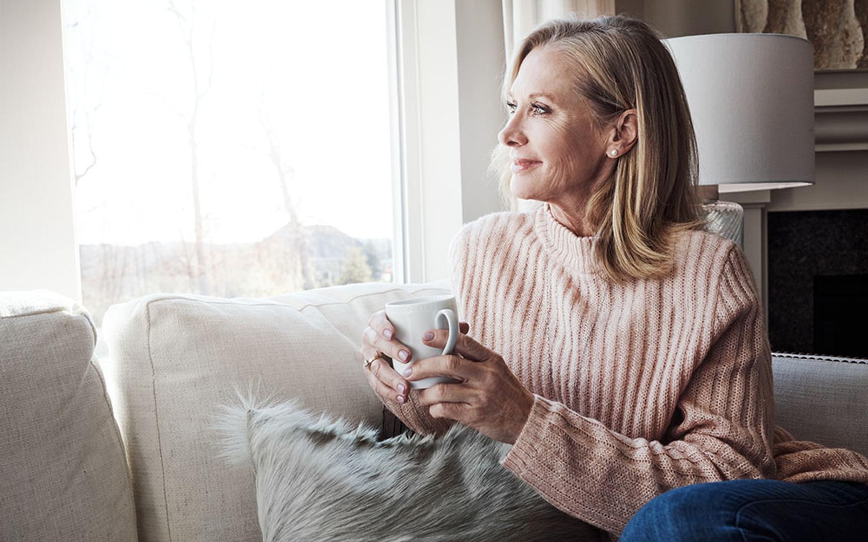 Dame som sitter i en sofa med en kopp te eller kaffe og ser ut av vinduet